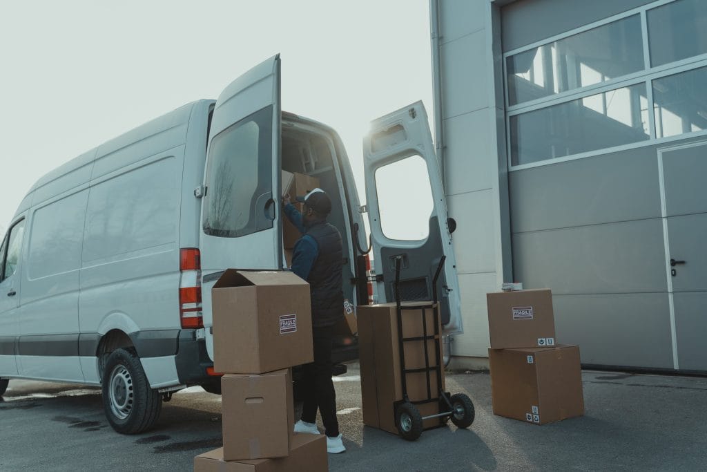 Courier loading cardboard boxes into a delivery van outside a warehouse.